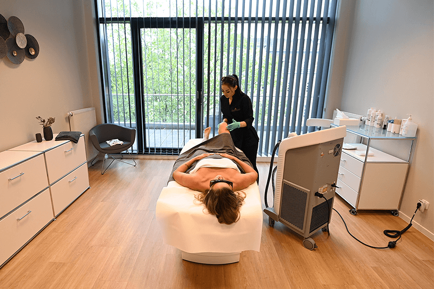 Behandlungsraum einer Schönheitsklinik mit moderner Einrichtung, in dem eine Kosmetikerin eine Patientin auf der Liege mit einem professionellen Lasergerät zur dauerhaften Haarentfernung behandelt.
