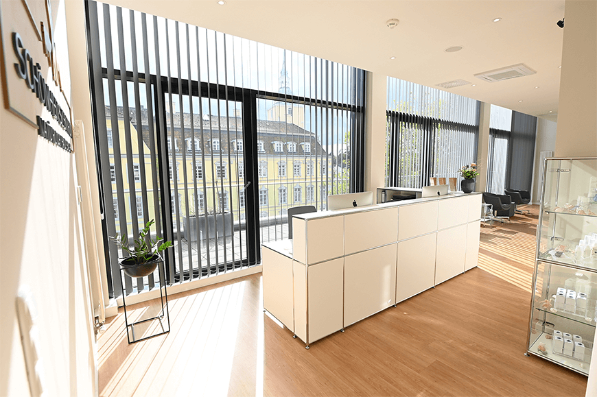 Heller, moderner Empfangsbereich einer Schönheitsklinik mit großer Fensterfront, vertikalen Lamellenjalousien, weißem Empfangstresen, Holzfußboden und Blick auf ein historisches Gebäude im Hintergrund.