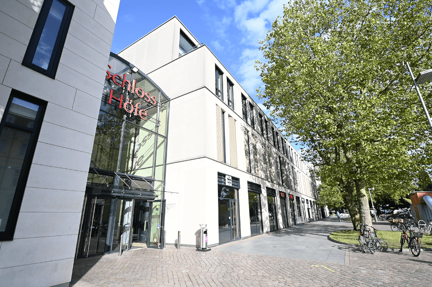 schoenheitszentrum-standort-oldenburg Eingangsbereich der Schloss Höfe in Oldenburg mit moderner Glasfassade, Einzelhandelsgeschäften im Erdgeschoss und Fahrradstellplätzen unter großen Bäumen an einem sonnigen Tag.