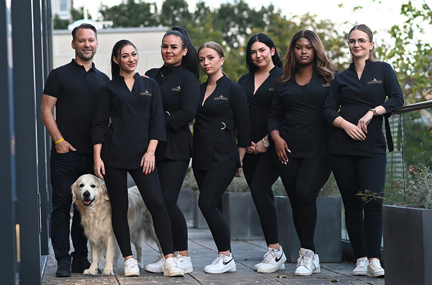 Teamfoto eines Schönheitszentrums: Sieben Mitarbeiterinnen und ein männlicher Kollege stehen gemeinsam mit einem Golden Retriever auf einer Terrasse, alle tragen schwarze Arbeitskleidung mit Logo.